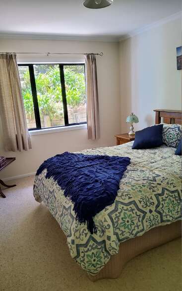 This cozy bedroom features a bright window with beige curtains, a floral-patterned bedspread, a dark blue throw, pillows, side table, and a vintage lamp.
