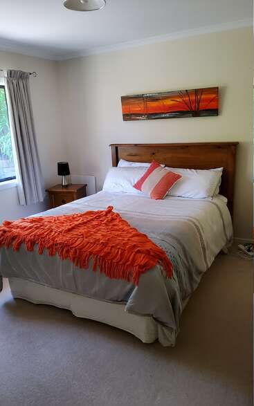 A cozy bedroom with a wooden bed, white bedding, and orange accents. Sunset artwork above bed, bedside table with black lamp, and bright natural light.