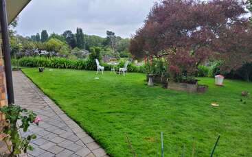 A lush green garden with a paved walkway, a flowering tree, white outdoor chairs and table, flowerbeds, and a peaceful, cloudy sky in the background.