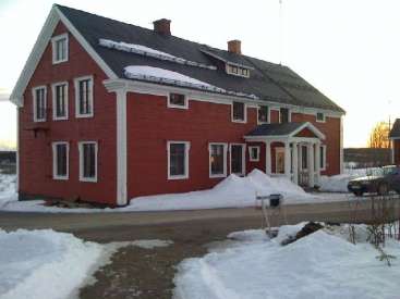 The image depicts a two-story red house with white trim, featuring a dark roof and chimney, situated in a snowy landscape with a driveway and mailbox in the foreground.