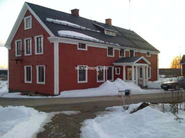 La imagen representa una gran casa roja con ribetes blancos y tejado oscuro, situada en un paisaje nevado con una carretera y un buzón en primer plano.
