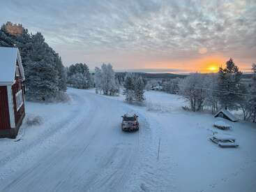 Un paisaje rural nevado con un coche rojo aparcado en una carretera sinuosa, una casa a la izquierda, árboles cubiertos de nieve y un amanecer resplandeciente.