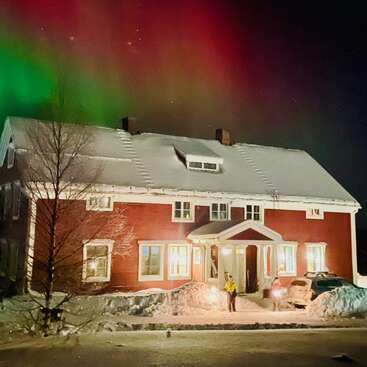 A red house covered in snow glows warmly at night. Above, vibrant green and red auroras light up the sky. One person stands outside, illuminated.