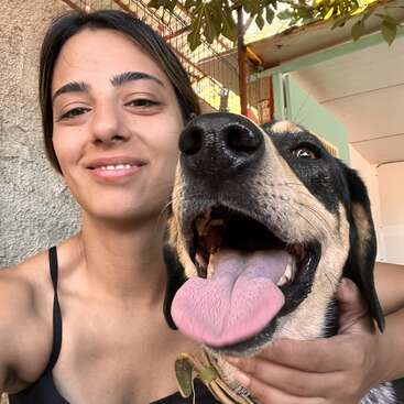 A young woman smiles while holding a happy dog with its tongue out. They appear to be outdoors, enjoying a sunny, cheerful moment together.