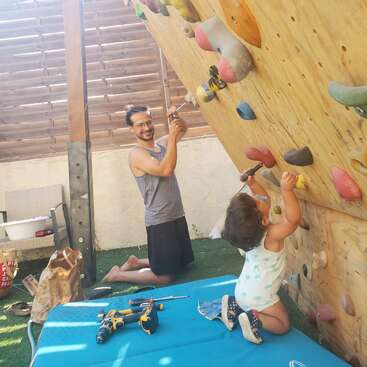 Un hombre y un niño pequeño trabajan juntos, utilizando herramientas para arreglar o construir un rocódromo al aire libre. Parecen felices y comprometidos, disfrutando de su actividad.