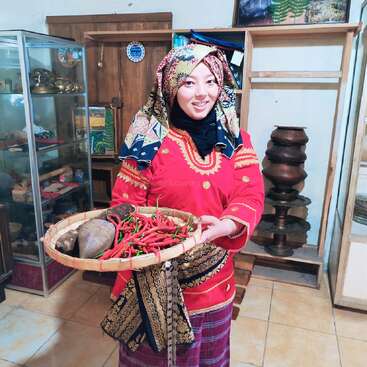 Eine Frau in farbenfroher traditioneller Kleidung lächelt, während sie ein geflochtenes Tablett mit roten Chilis und Knollen in der Hand hält, während sie in einem Raum mit kulturellen Artefakten steht.
