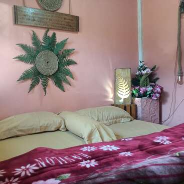 Ein gemütliches Schlafzimmer mit einer rosafarbenen Wand, beigen Kissen, einer roten Blumendecke, dekorativer Wandkunst, Farnen, einer Holzlampe und einem Strauß Kunstblumen.