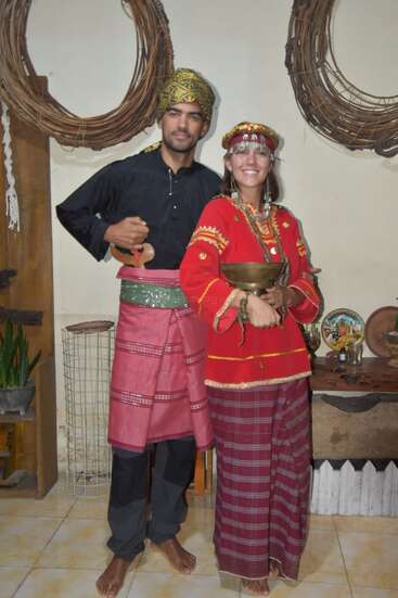 Ein Mann und eine Frau in traditioneller indonesischer Kleidung posieren in einem Haus und lächeln. Der Mann trägt ein schwarzes Hemd, einen rosafarbenen Sarong und eine goldene Kopfbedeckung, die Frau trägt ein rotes Gewand.