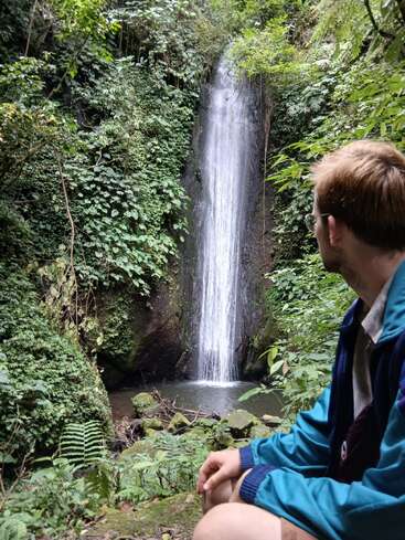 Ein Mann in einer blauen Jacke sitzt in einem üppigen, grünen Wald und bewundert friedlich einen schmalen Wasserfall, der in ein kleines, von Laub umgebenes Becken stürzt.
