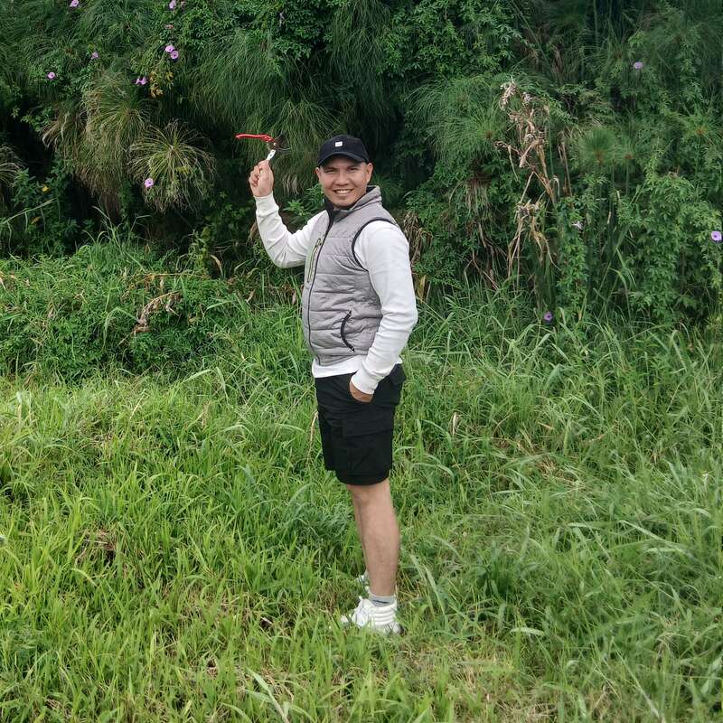 Um homem sorridente está sobre uma grama verde exuberante, segurando uma tesoura de jardinagem. Ele usa boné, colete cinza, camisa branca, bermuda preta e tênis branco, com vegetação atrás.