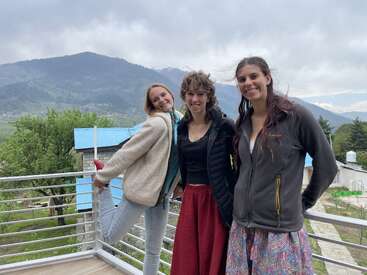 Trois femmes se tiennent sur un balcon, souriantes, avec un magnifique paysage de montagne et un ciel nuageux en arrière-plan. Elles semblent heureuses et détendues, profitant de la vue panoramique.