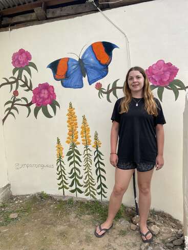Une jeune femme se tient à côté d'une fresque murale représentant un grand papillon bleu et orange, des fleurs roses et de grandes fleurs sauvages jaunes, sur un mur extérieur blanc.