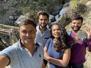 Quatre amis posent pour un selfie joyeux en plein air. Derrière eux, un terrain rocheux, de la verdure et un ruisseau qui coule forment un magnifique décor naturel, ensoleillé et propice à l'aventure.