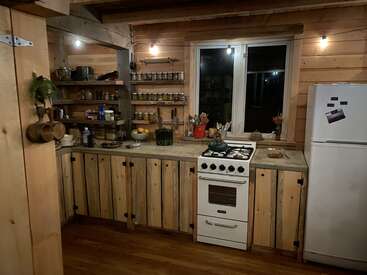 Cette cuisine rustique et chaleureuse présente des murs et des armoires en bois, des étagères ouvertes avec des bocaux, une cuisinière blanche, un réfrigérateur, une fenêtre et un éclairage chaleureux qui créent une atmosphère accueillante.