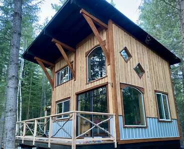 Une cabane moderne en bois nichée dans une forêt, avec de grandes fenêtres cintrées, une terrasse enveloppante, des accents géométriques et un toit haut et en surplomb pour faire de l'ombre.