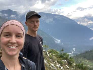 Uma mulher sorridente e um homem estão em uma trilha na montanha, cercados por uma vegetação exuberante e montanhas dramáticas e enevoadas sob um céu nublado. Momento cênico e aventureiro.