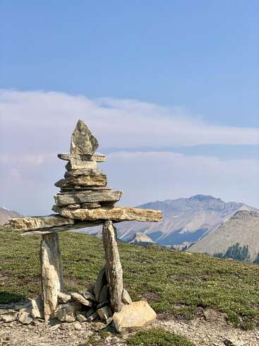 Un Inukshuk soigneusement équilibré, fait de roches plates, se dresse sur une crête montagneuse herbeuse, avec derrière lui des pics escarpés lointains et un ciel bleu clair et lumineux.