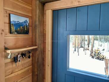 Un intérieur de cabane en bois confortable avec une porte bleue, une fenêtre donnant sur les bois enneigés, des crochets pour les clés, une petite étagère et un paysage de montagne sur le mur.