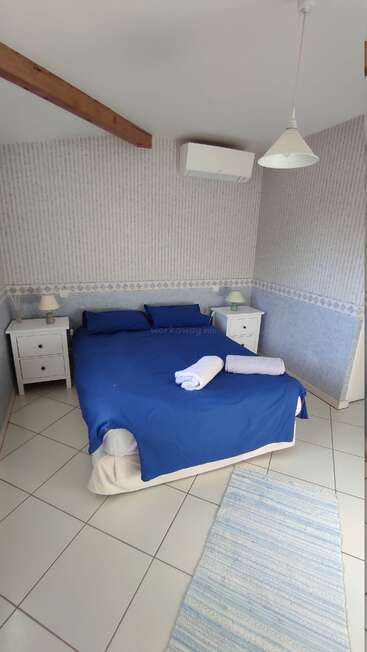 A imagem mostra um quarto sereno com uma cama azul, mesinhas de cabeceira brancas e uma luminária suspensa, com um tapete azul e branco em um piso de azulejos.