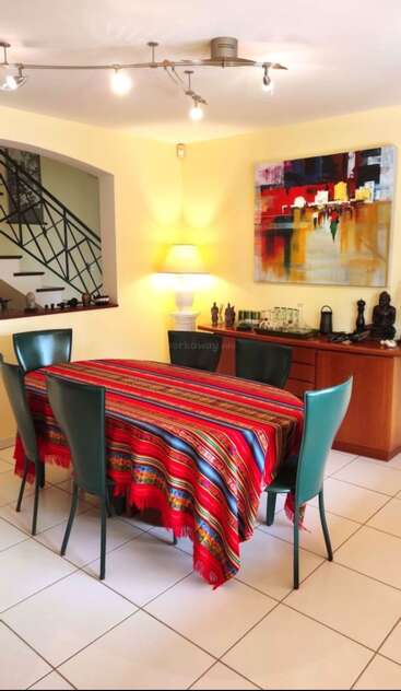 Uma sala de jantar aconchegante apresenta uma toalha de mesa vibrante e listrada, cadeiras verdes, arte abstrata moderna, luz suave de abajur, um aparador com ornamentos e vista para a escada.