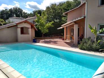 Uma piscina azul cintilante fica ao lado de uma casa bege, cercada por vegetação e árvores. O sol brilha intensamente, projetando sombras no pátio e nas áreas de relaxamento.