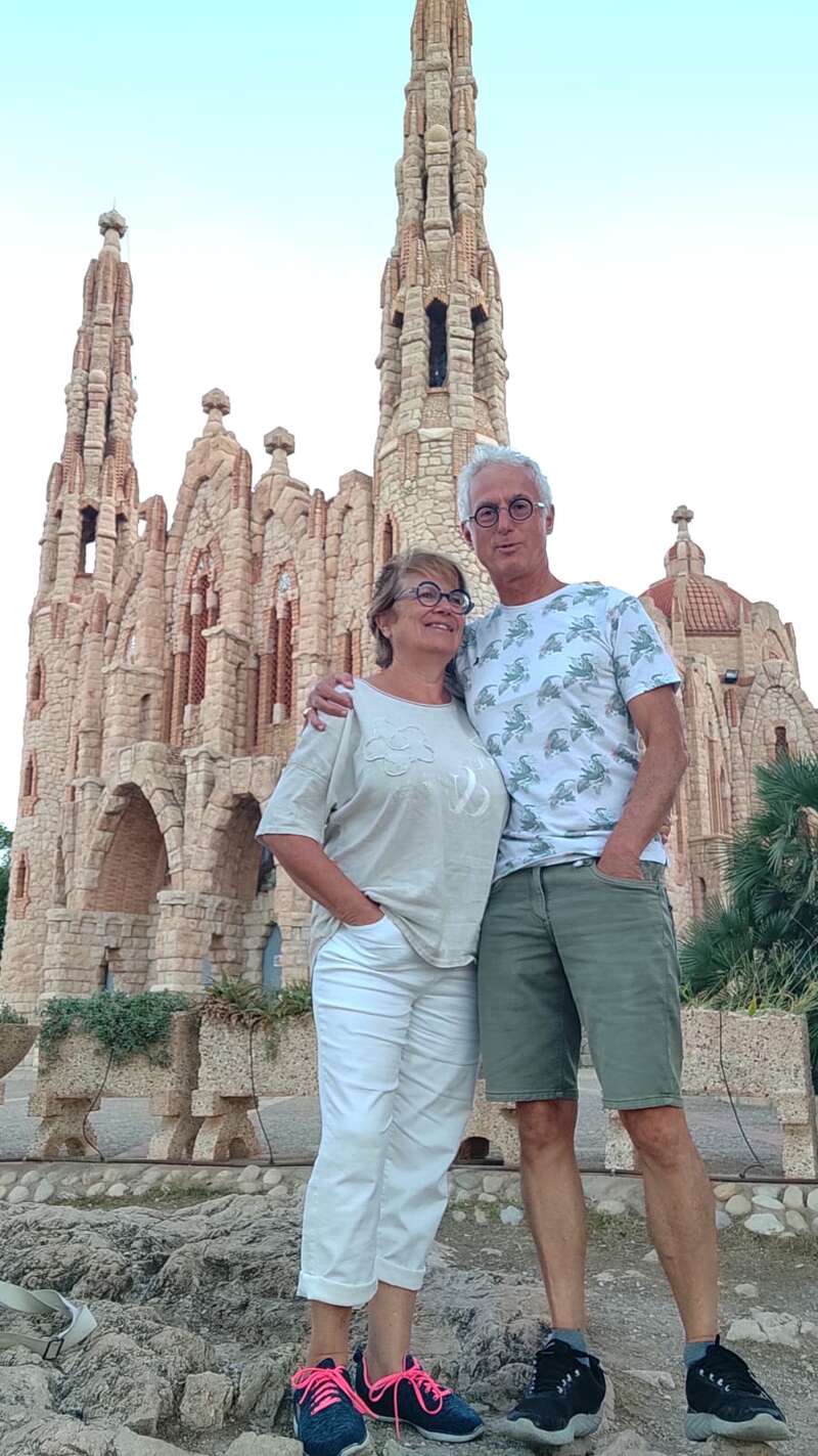 A imagem mostra um casal de meia-idade em frente a um grande edifício de pedra, com a mulher à esquerda e o homem à direita, ambos vestindo roupas casuais.