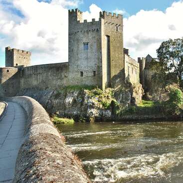 La imagen representa un castillo con foso, con un muro de piedra y un puente, rodeado por una masa de agua, con una zona de césped en primer plano.