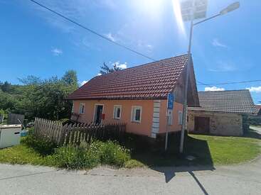 Una pequeña casa rosa con tejado de tejas rojas se alza junto a una calle tranquila, rodeada de vegetación, una valla de madera y un cielo azul y despejado sobre ella.