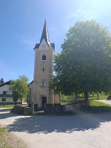 Una pequeña iglesia con un campanario puntiagudo se alza junto a un gran árbol, bajo un cielo azul y despejado, rodeada de vegetación y de tranquilas casas de pueblo. Un ambiente apacible.