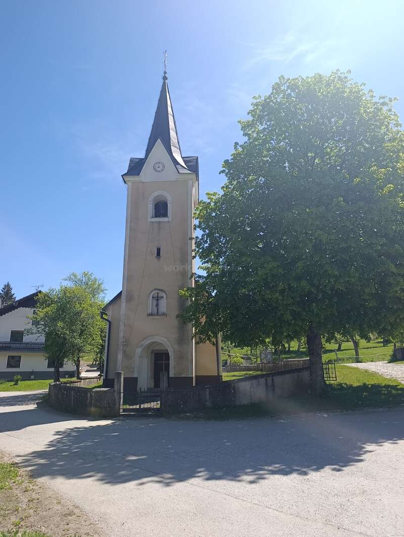 Una pequeña iglesia con un campanario puntiagudo se alza junto a un gran árbol, bajo un cielo azul y despejado, rodeada de vegetación y de tranquilas casas de pueblo. Un ambiente apacible.