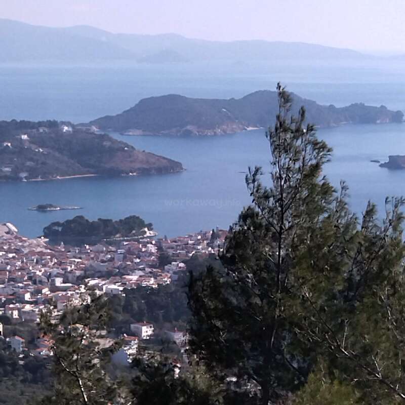 Cette image montre une ville côtière pittoresque avec des bâtiments blancs, entourée d'arbres verdoyants, surplombant une mer bleue sereine et plusieurs petites îles.