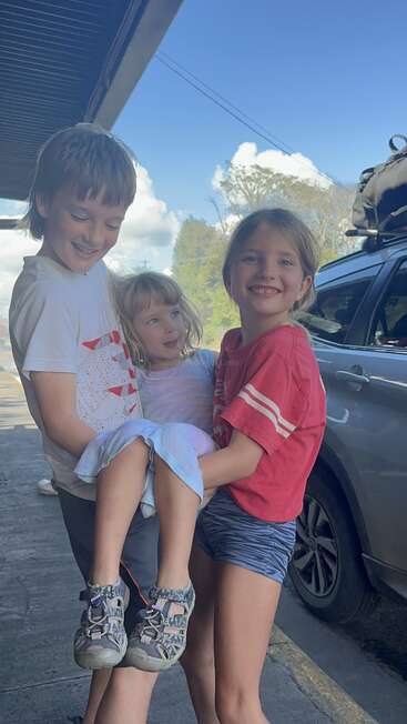 Três crianças sorridentes posam juntas ao ar livre. Duas crianças mais velhas, um menino e uma menina, seguram alegremente uma menina mais nova. Um carro e um céu azul ao fundo.