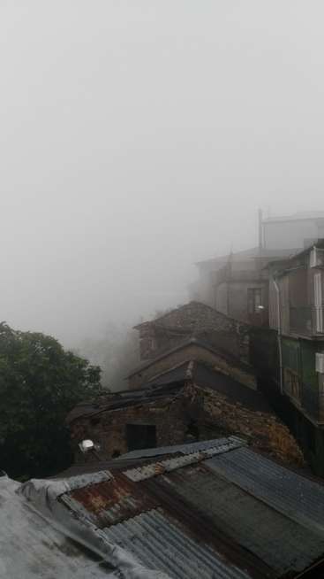 La imagen representa una escena brumosa de edificios viejos con tejados oxidados, en la que aparece un árbol a la izquierda y una estructura de piedra en el centro.