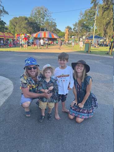 Une famille de quatre personnes souriantes pose en plein air dans un parc d'attractions animé, avec en arrière-plan des tentes colorées, des manèges et des gens profitant d'une journée ensoleillée et divertissante.