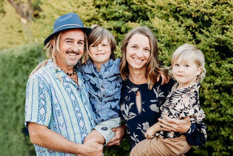 Une famille heureuse de quatre personnes pose en plein air. Le père porte un chapeau et une chemise à motifs, la mère sourit chaleureusement, les deux fils portent des chemises ludiques, et une végétation luxuriante se trouve derrière eux.