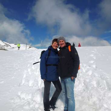 The image depicts a couple standing in the snow, with a man and woman embracing, wearing winter attire, set against a backdrop of a snowy landscape and a blue sky with clouds.