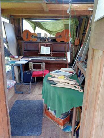 Una acogedora y rústica sala de música con un piano, dos guitarras encima, partituras esparcidas, una mesa cubierta de verde, sillas, paredes de madera y varios instrumentos musicales.