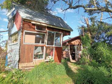 Una extravagante casa de madera hecha a mano con grandes ventanas desiguales está rodeada de exuberante vegetación, junto a una acogedora cabaña bajo un brillante cielo azul.
