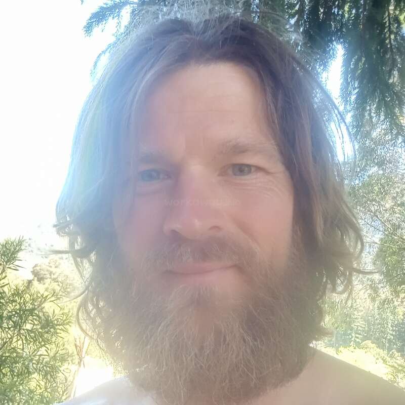 Un hombre con barba y pelo largo está de pie al aire libre, sonriendo suavemente a la cámara. La luz del sol se filtra a través del verde follaje que hay detrás de él, creando un apacible fondo natural.