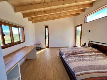 Este quarto luminoso e minimalista tem piso de madeira, vigas à vista no teto, uma cama grande, uma escrivaninha embutida, janelas amplas e um estilo moderno e simples, com muita luz natural e espaço.