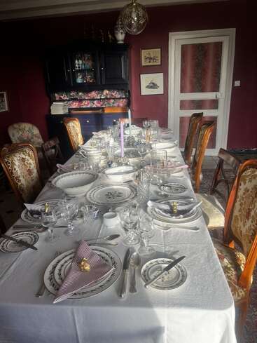The image depicts a formal dining room with a long table set for a meal, featuring a white tablecloth, plates, glasses, silverware, and napkins, surrounded by chairs and a hutch.