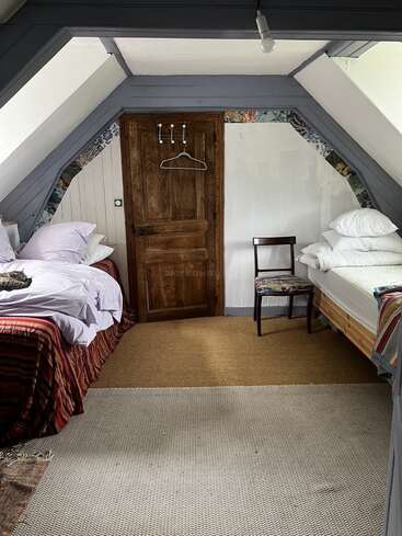 The image depicts a cozy attic bedroom with sloping ceilings, featuring two beds, a wooden door, and a chair, exuding a warm and inviting atmosphere.