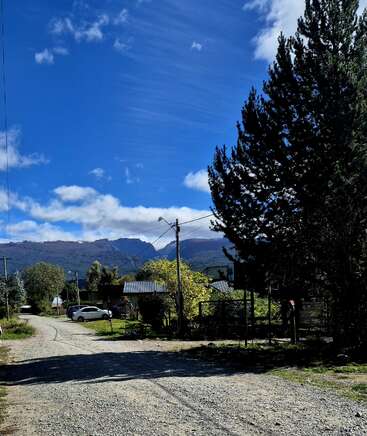 Uma estrada de terra sinuosa passa por casas e carros estacionados, cercada por árvores e montanhas sob um céu azul vibrante salpicado de nuvens brancas e fofas. Uma cena tranquila.