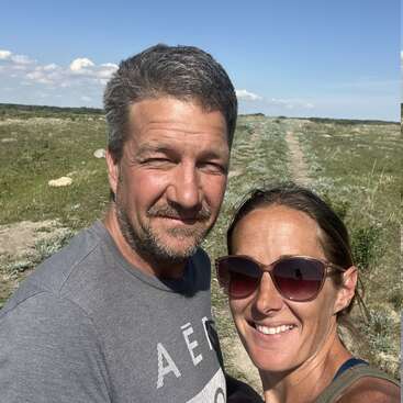 A smiling man and woman take a selfie outdoors on a sunny day, standing in a grassy field under a blue sky with scattered clouds.