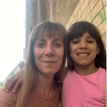 Uma mulher e uma jovem, ambas sorridentes, posam juntas ao ar livre ao lado de um muro. A garota está usando uma camiseta rosa e tem o braço em volta da mulher.