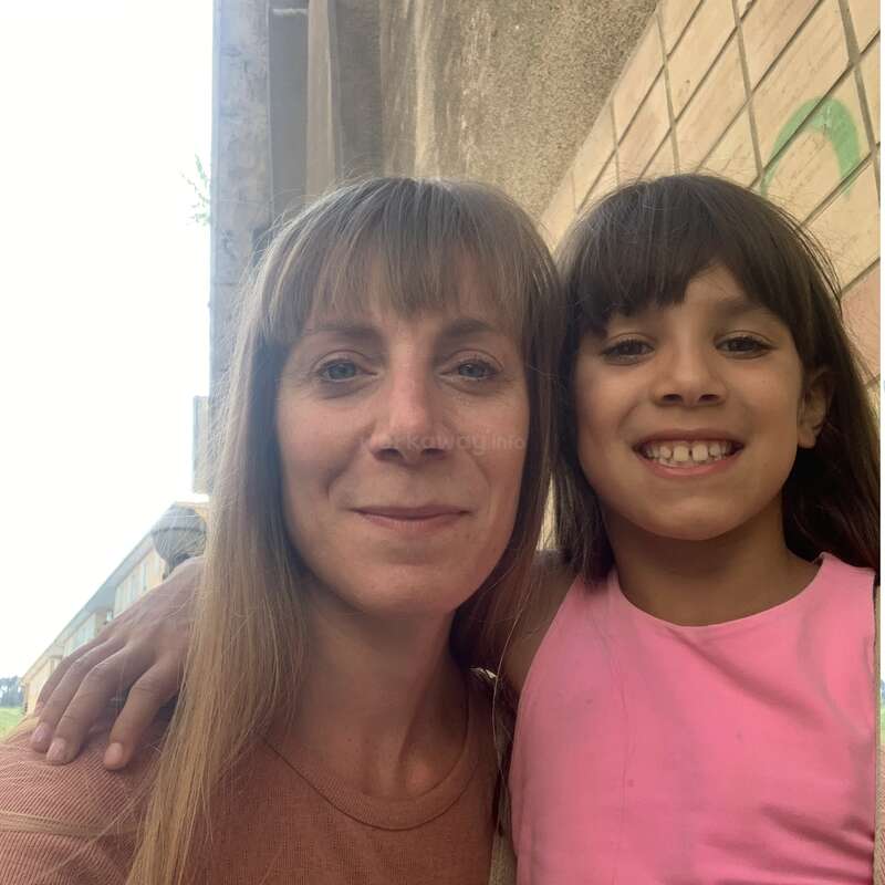 A woman and a young girl, both smiling, pose together outdoors beside a wall. The girl is wearing a pink shirt and has her arm around the woman.