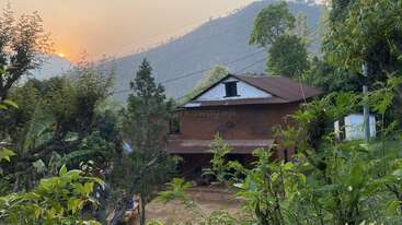 Uma casa rústica de tijolos está aninhada entre árvores verdejantes e colinas ao fundo. O sol está se pondo, lançando um brilho quente e pacífico.