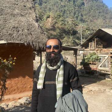A bearded man wearing sunglasses, a black sweater, and a striped scarf stands outdoors near rustic huts, with a green mountainous background and sunshine illuminating the scene.