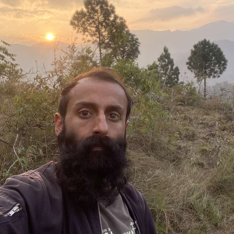 Um homem barbudo tira uma selfie em uma paisagem gramada e montanhosa ao pôr do sol. Você pode ver pinheiros e montanhas ao fundo sob um céu brilhante.