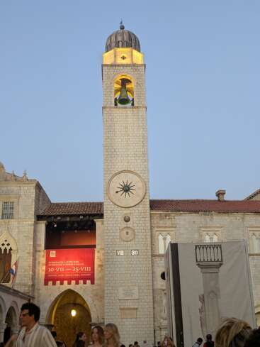 Uma torre de relógio alta e iluminada com um sino fica em uma praça histórica ao anoitecer. As pessoas caminham abaixo, e faixas anunciam o Festival de Verão de Dubrovnik.